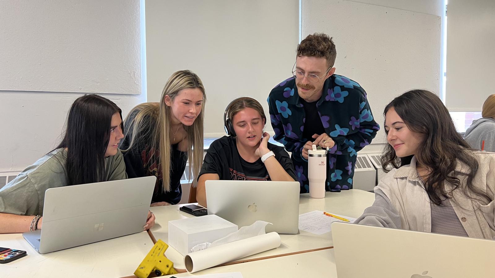 Four students seated looking at a peer's work on a laptop with their instructor standing and participating in discussion. 